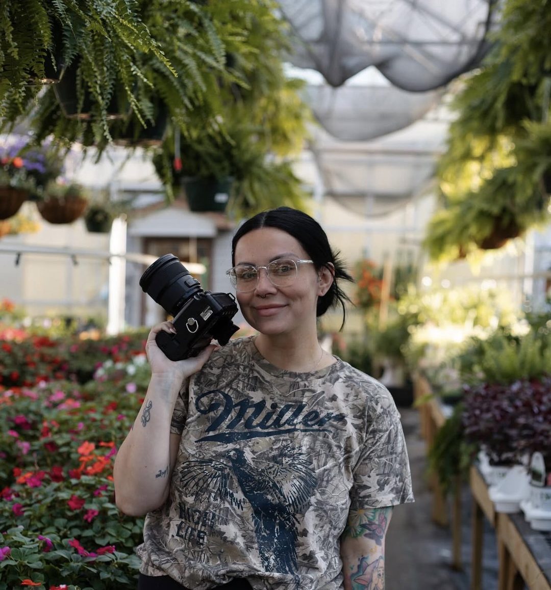 Taylor, camera in hand, smiling in a greenhouse full of flowers