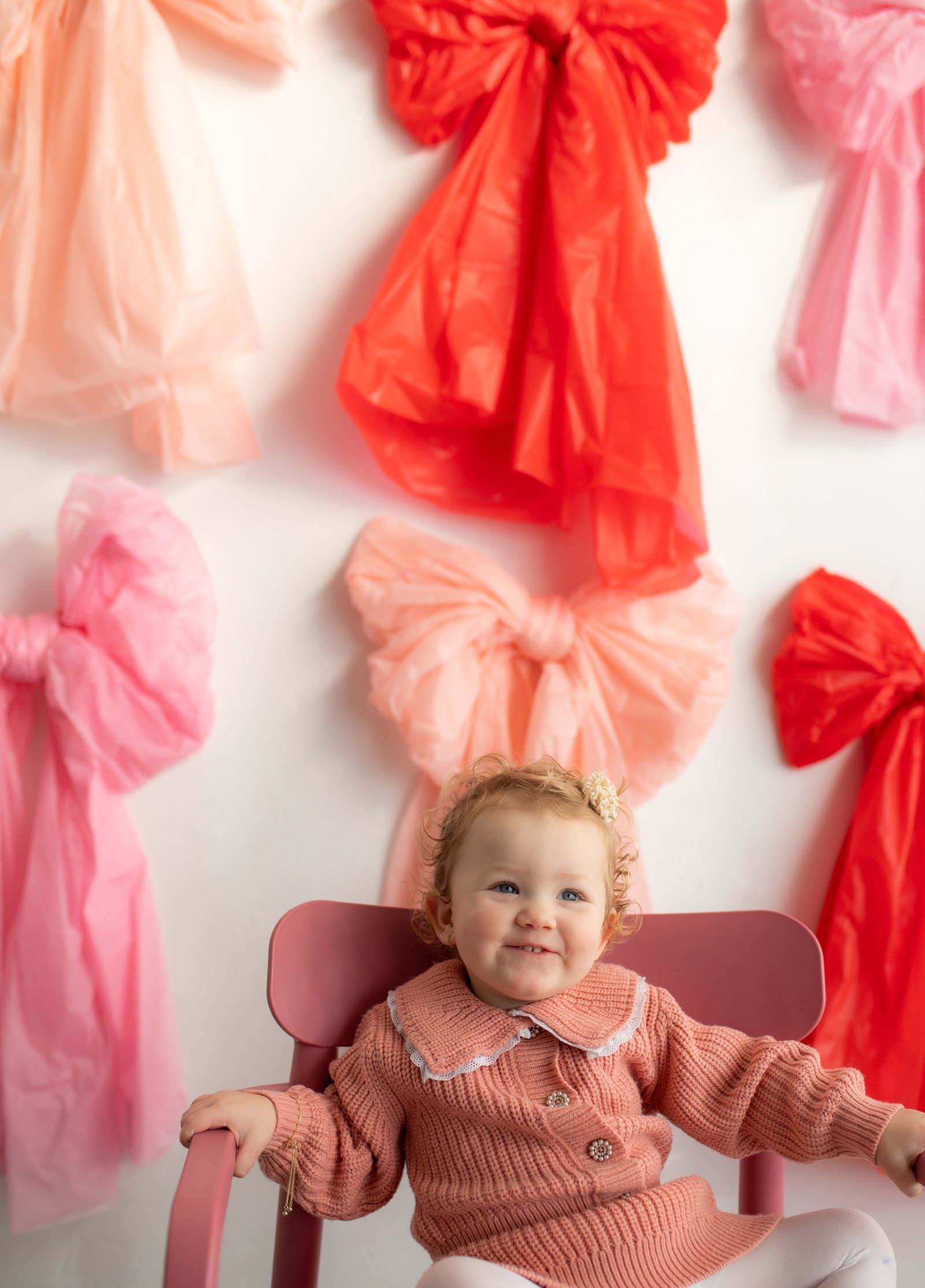 A smiling toddler in a pink sweater seated in front of a wall of pink and red tulle bows