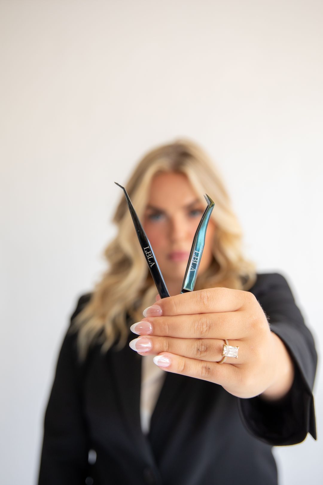 A lash artist in a black blazer holding two lash tweezers out toward the camera