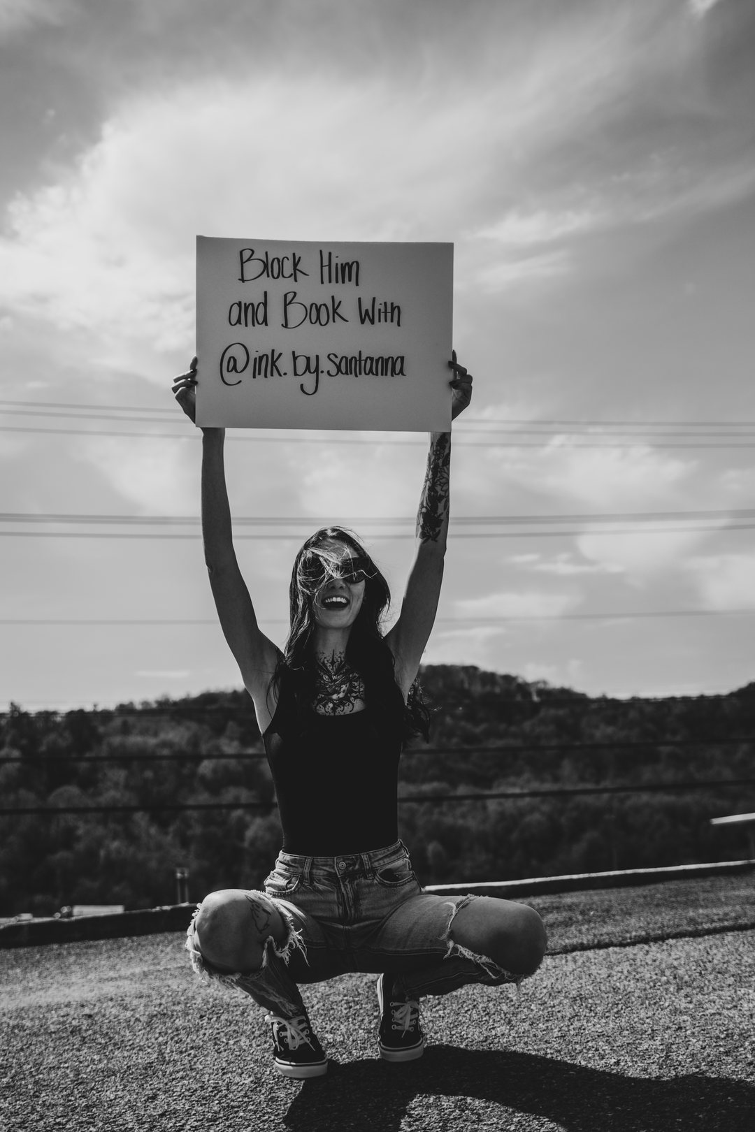 Black and white photo of a tattoo artist holding a hand-lettered sign above her head