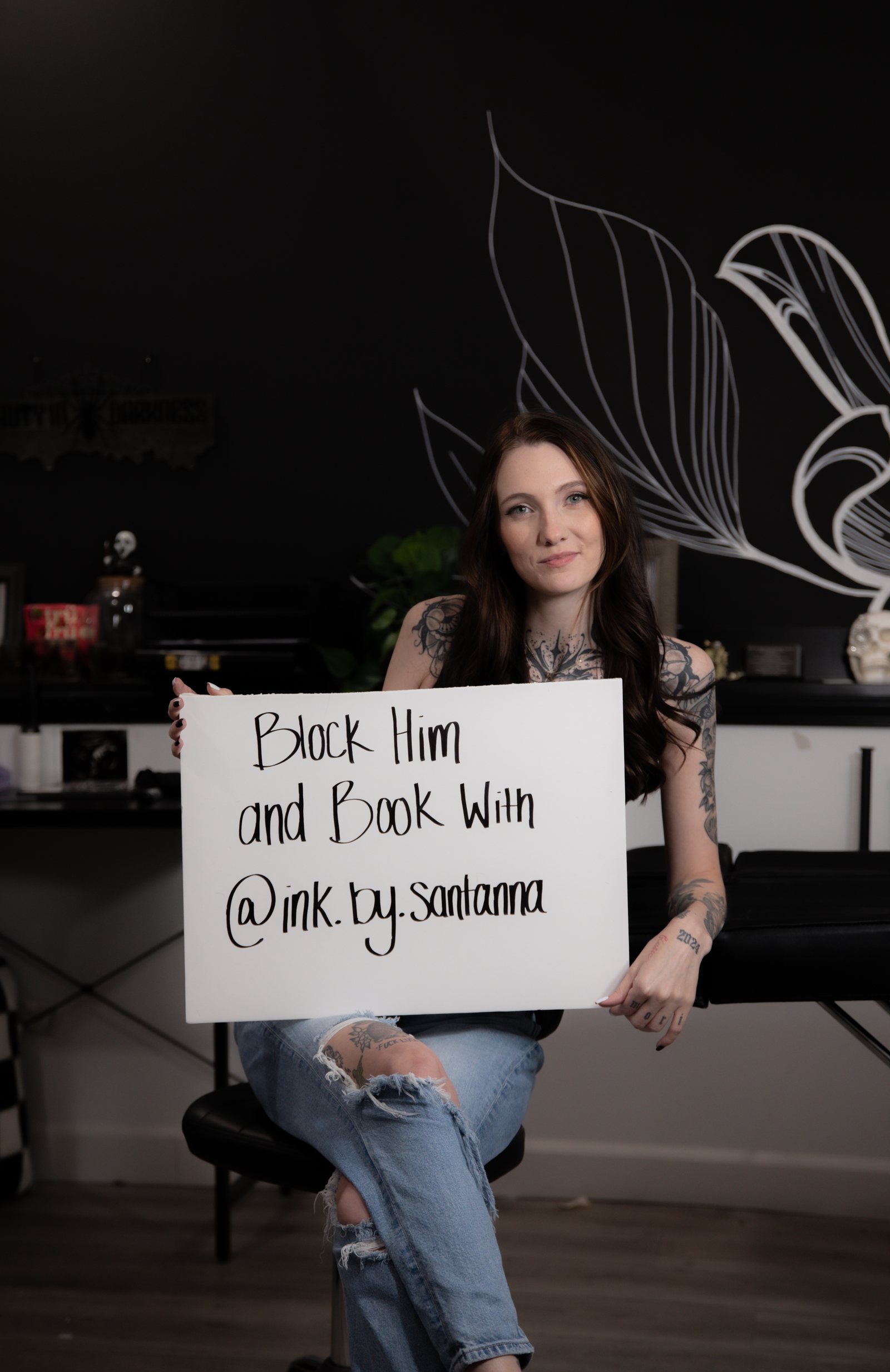 A tattoo artist seated in her studio holding a hand-lettered booking sign
