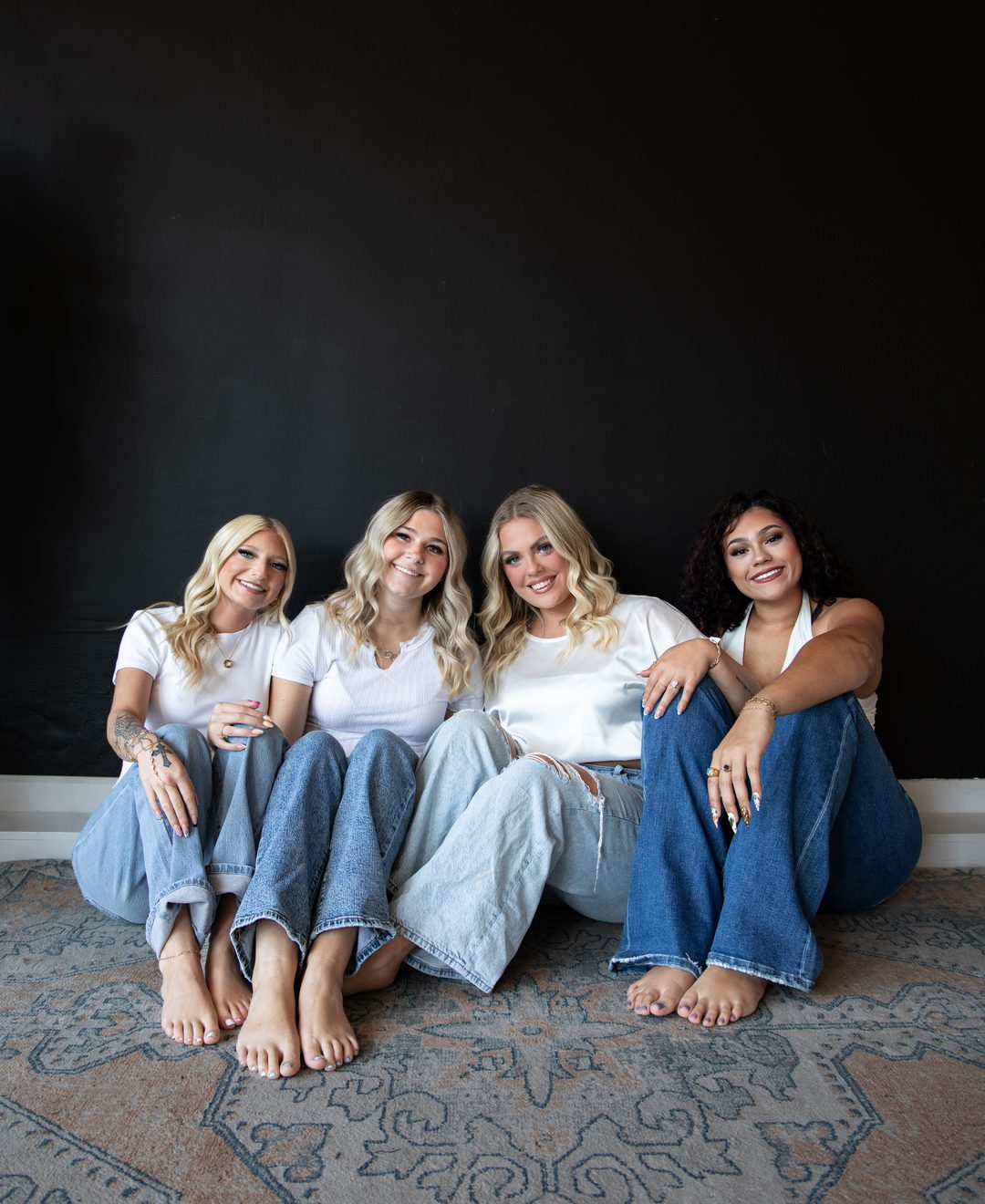 Four women in white tops and jeans sitting shoulder to shoulder against a black wall