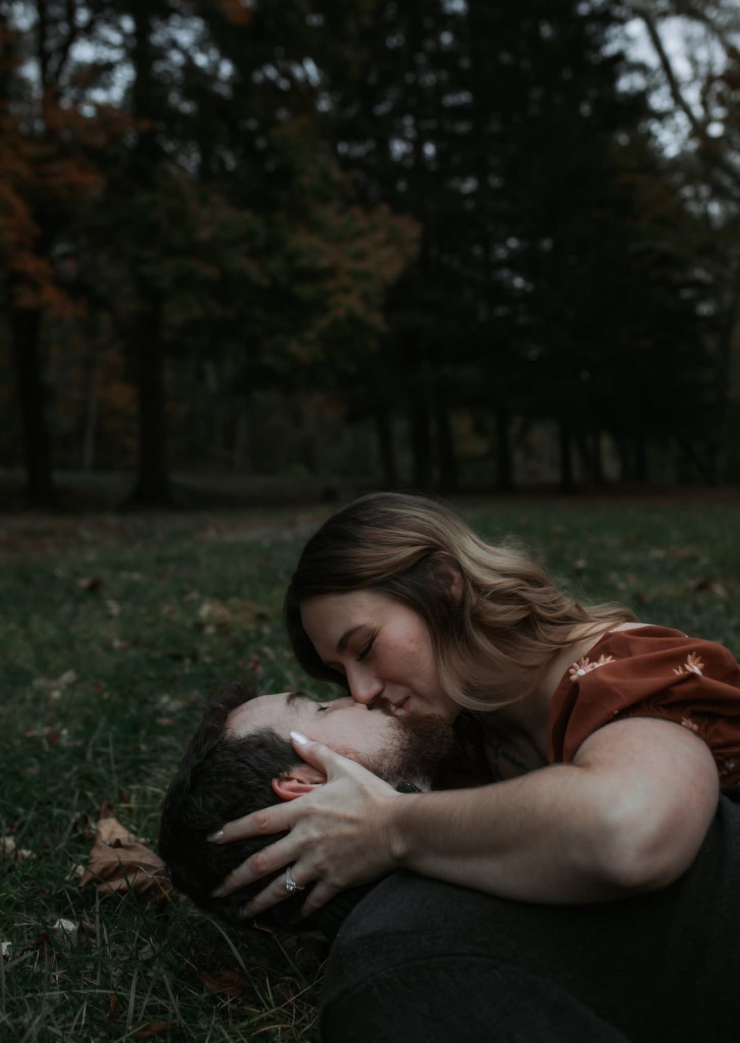 A couple lying in the grass at dusk, she leans down to kiss him