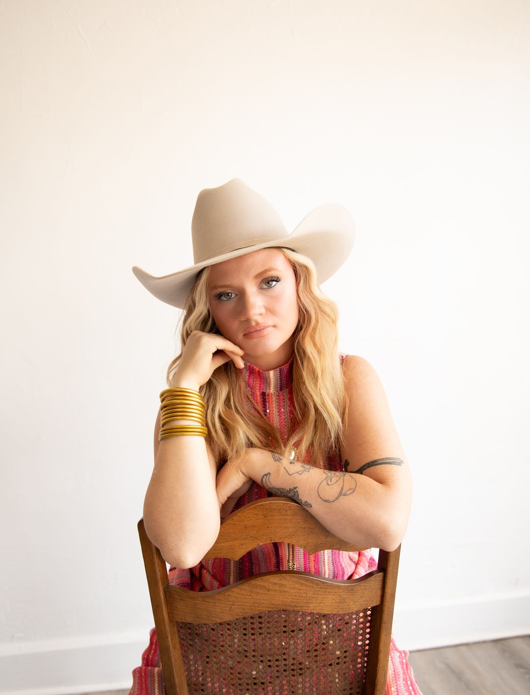 A woman in a cream cowboy hat and pink knit dress leaning on the back of a wooden chair