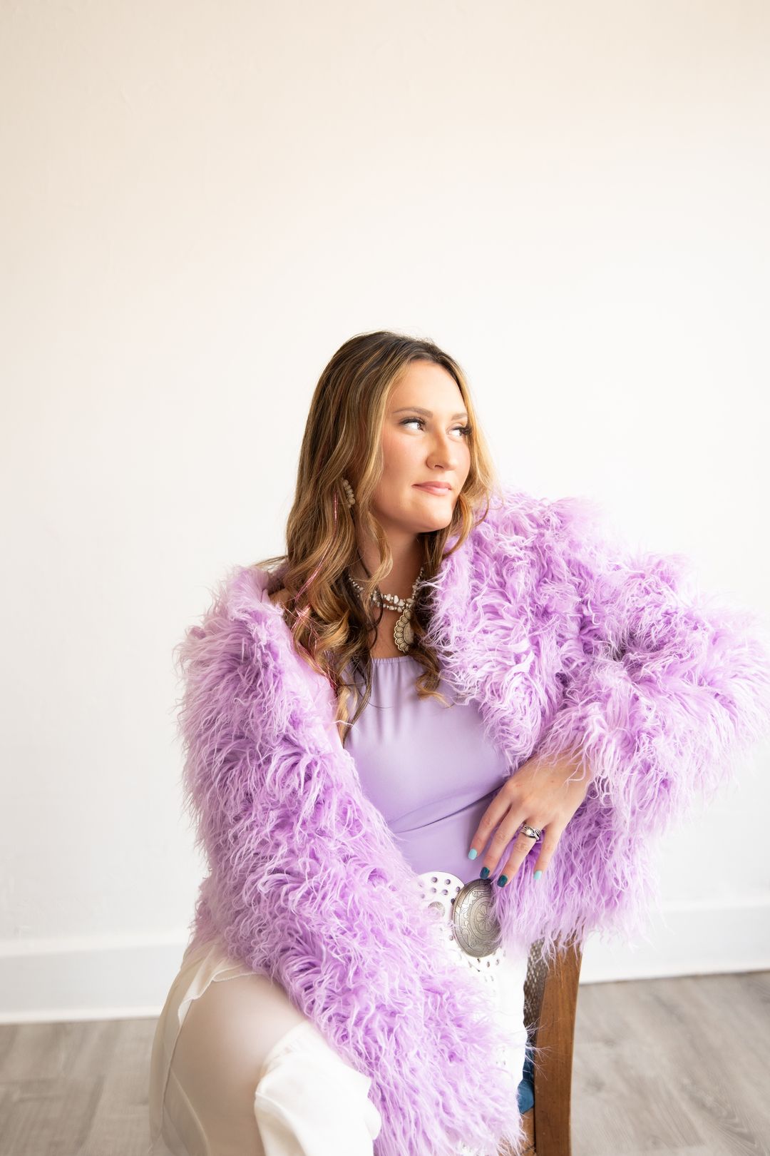A woman in a fluffy lavender jacket and concho belt seated in a vintage chair
