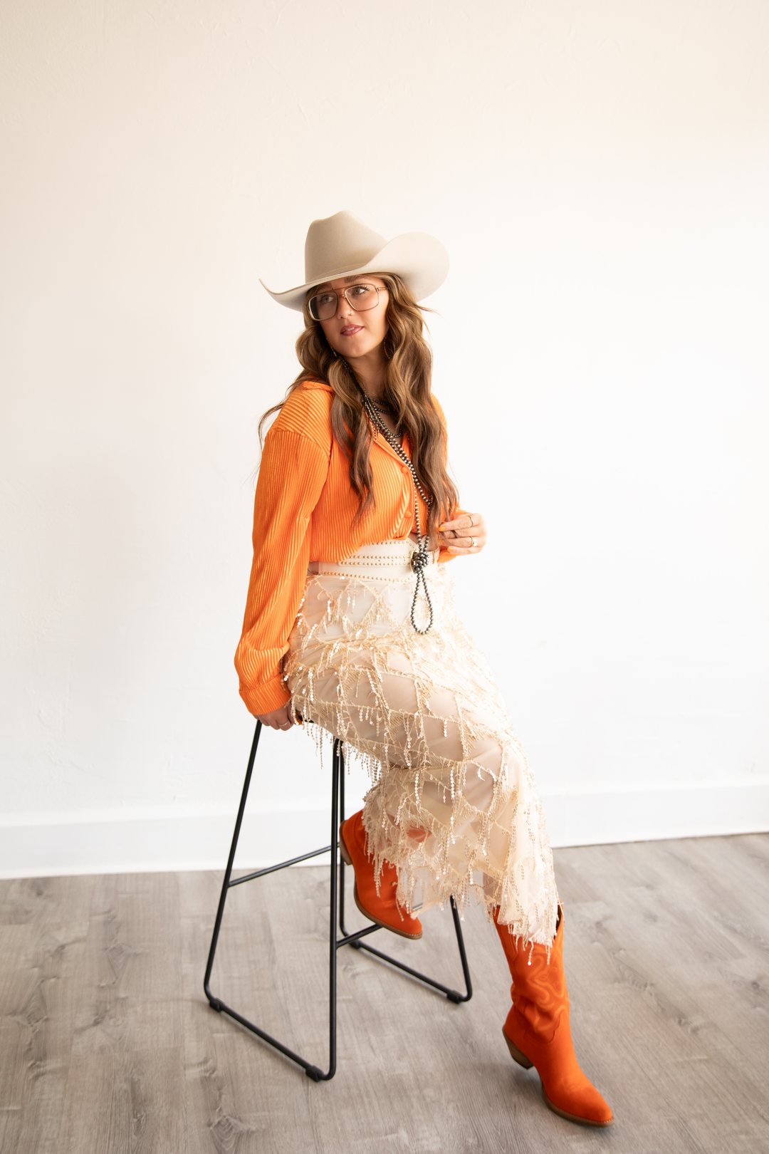 A woman in a cowboy hat and orange boots seated on a stool