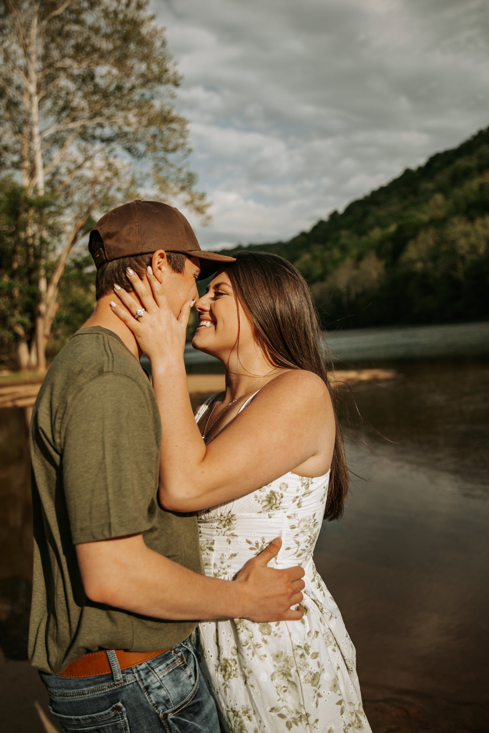 An engaged couple laughing forehead to forehead by the river, her ring catching the light