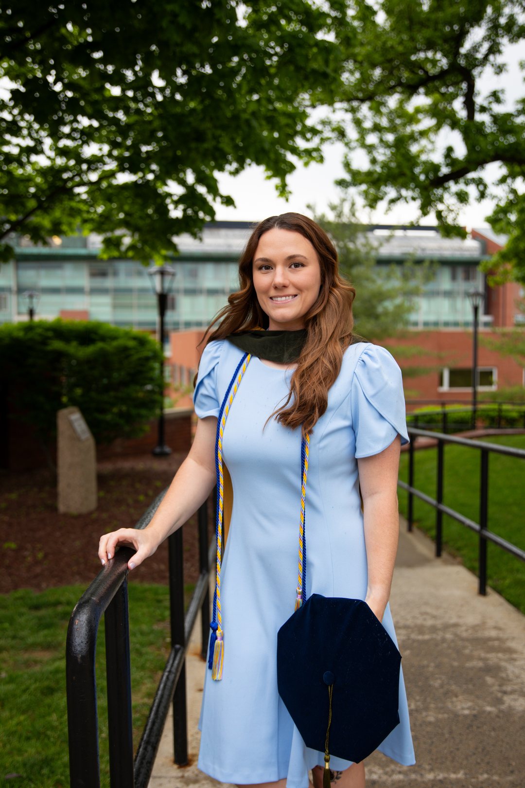 A graduate in a light blue dress holding her doctoral cap on campus