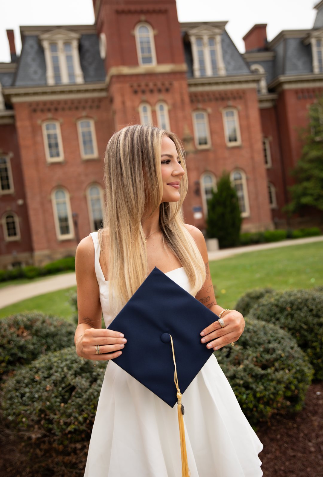 A graduate in a white dress holding her cap in front of Woodburn Hall