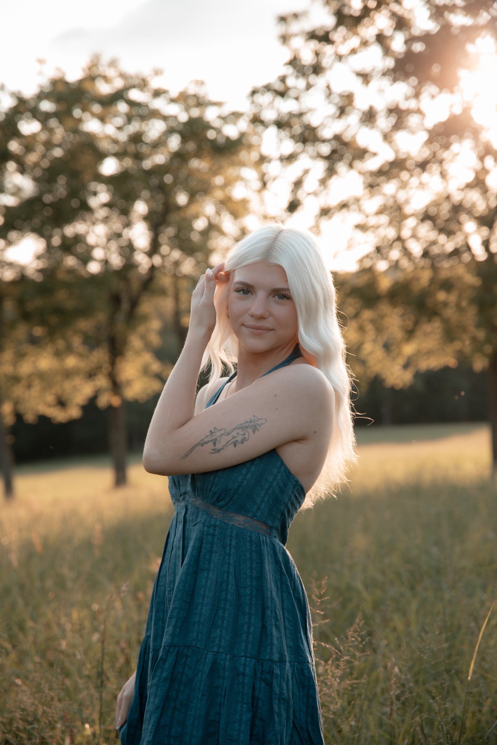 A senior in a blue dress backlit by golden hour sun in a meadow