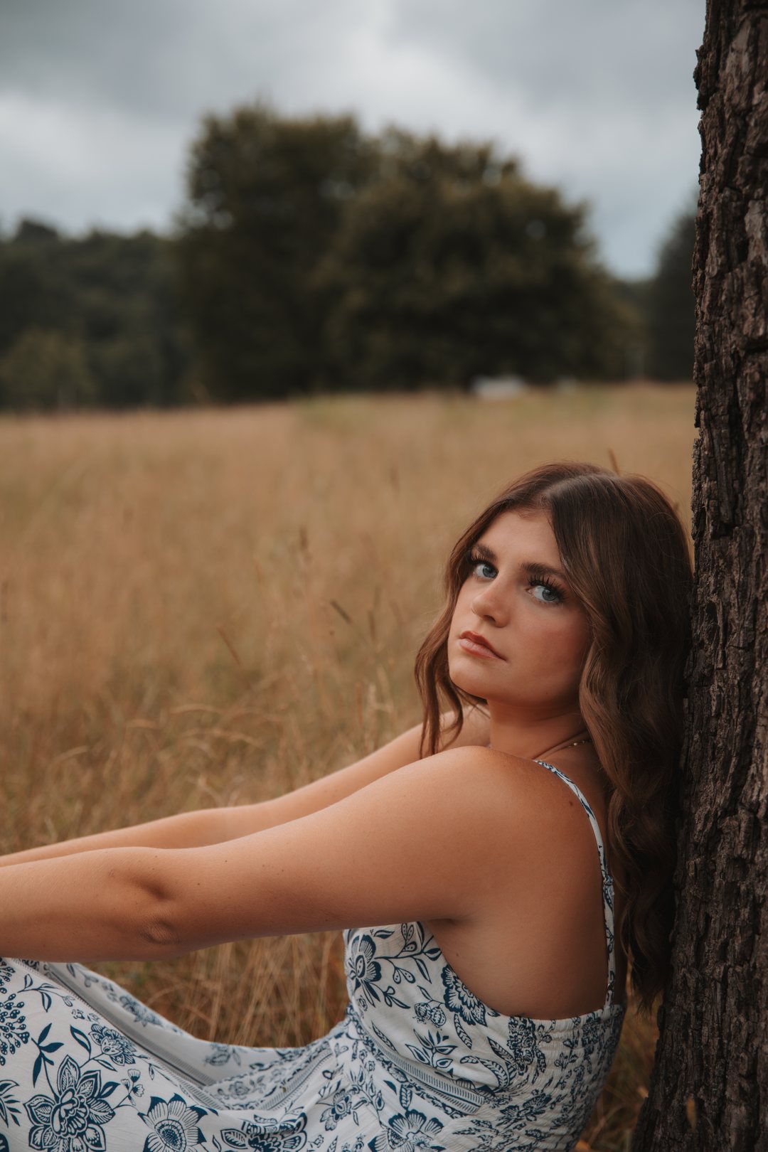 A senior in a blue floral dress leaning against a tree in an open field