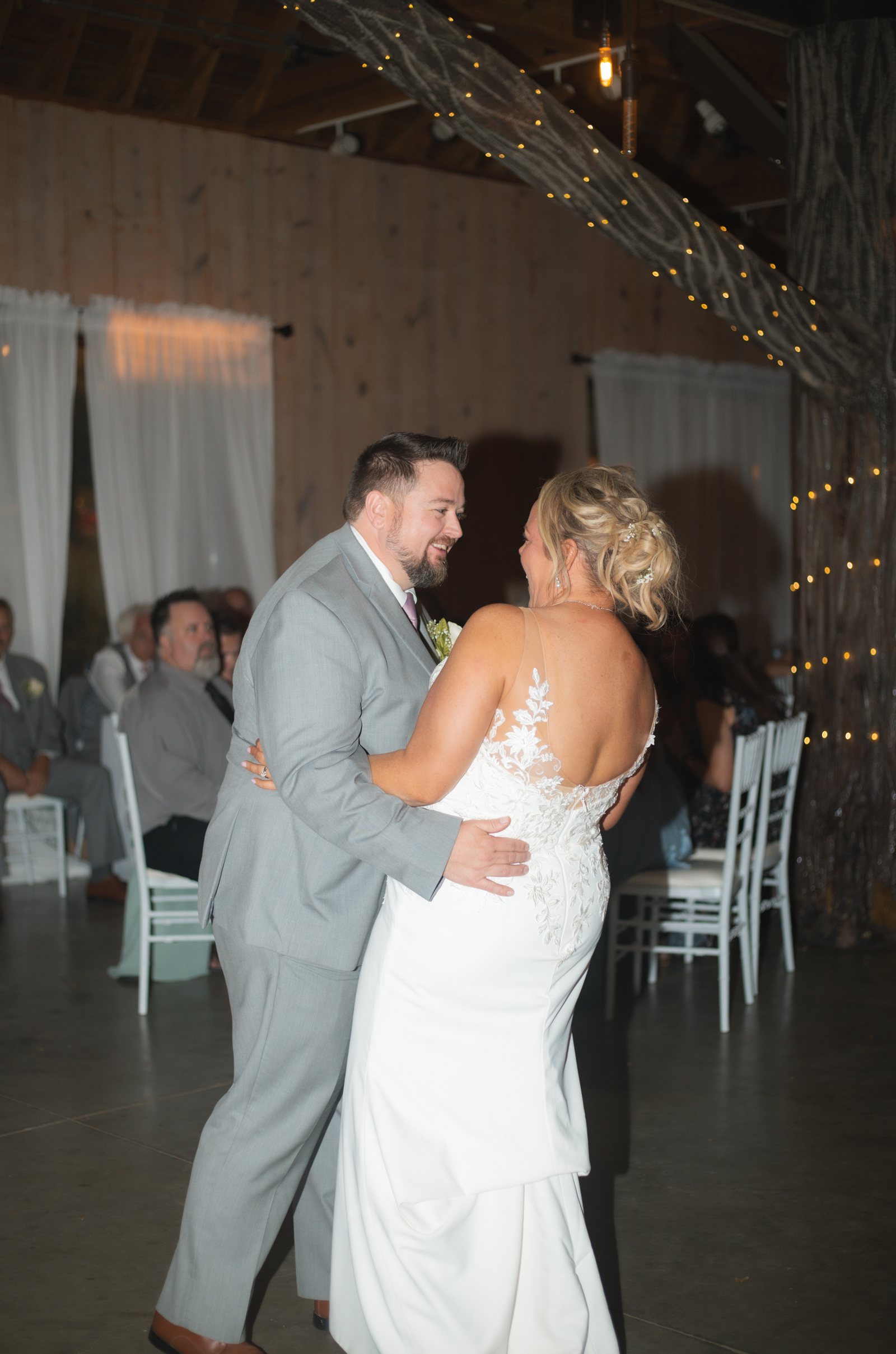 A bride and groom during their first dance under string lights in a barn