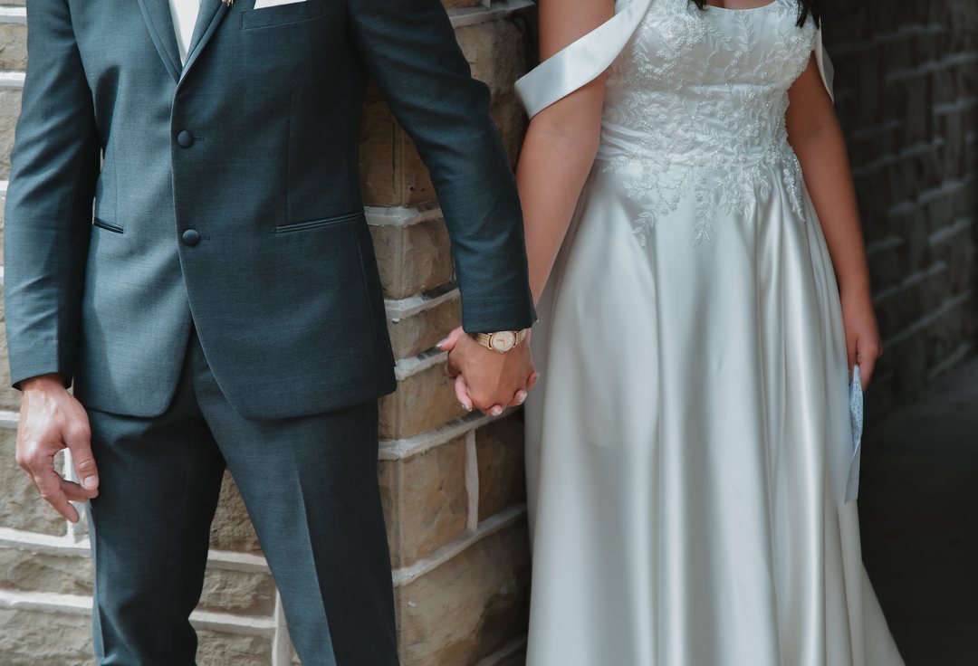 A bride and groom holding hands, detail of the dress and suit